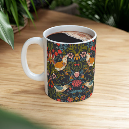 A white ceramic mug filled with black coffee sits on a light wood table, featuring a detailed botanical and bird pattern in dark green, orange, pink, and blue. Green plants are visible in the blurred background.