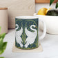 A ceramic mug with a design of two white swans on a green and blue background, inspired by the art of William Morris.