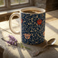 A ceramic mug filled with coffee sits on a rustic wooden table draped with a beige cloth, alongside a silver spoon and a small sprig of lavender. The mug features a navy blue background with a cream-colored floral vine pattern and red-orange flower accents.