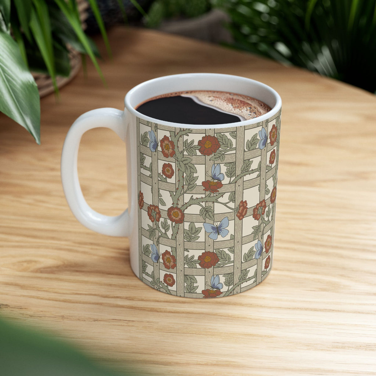 A white ceramic coffee mug with a vintage-style floral and butterfly pattern, filled with black coffee and resting on a light wooden table with green plants in the background.