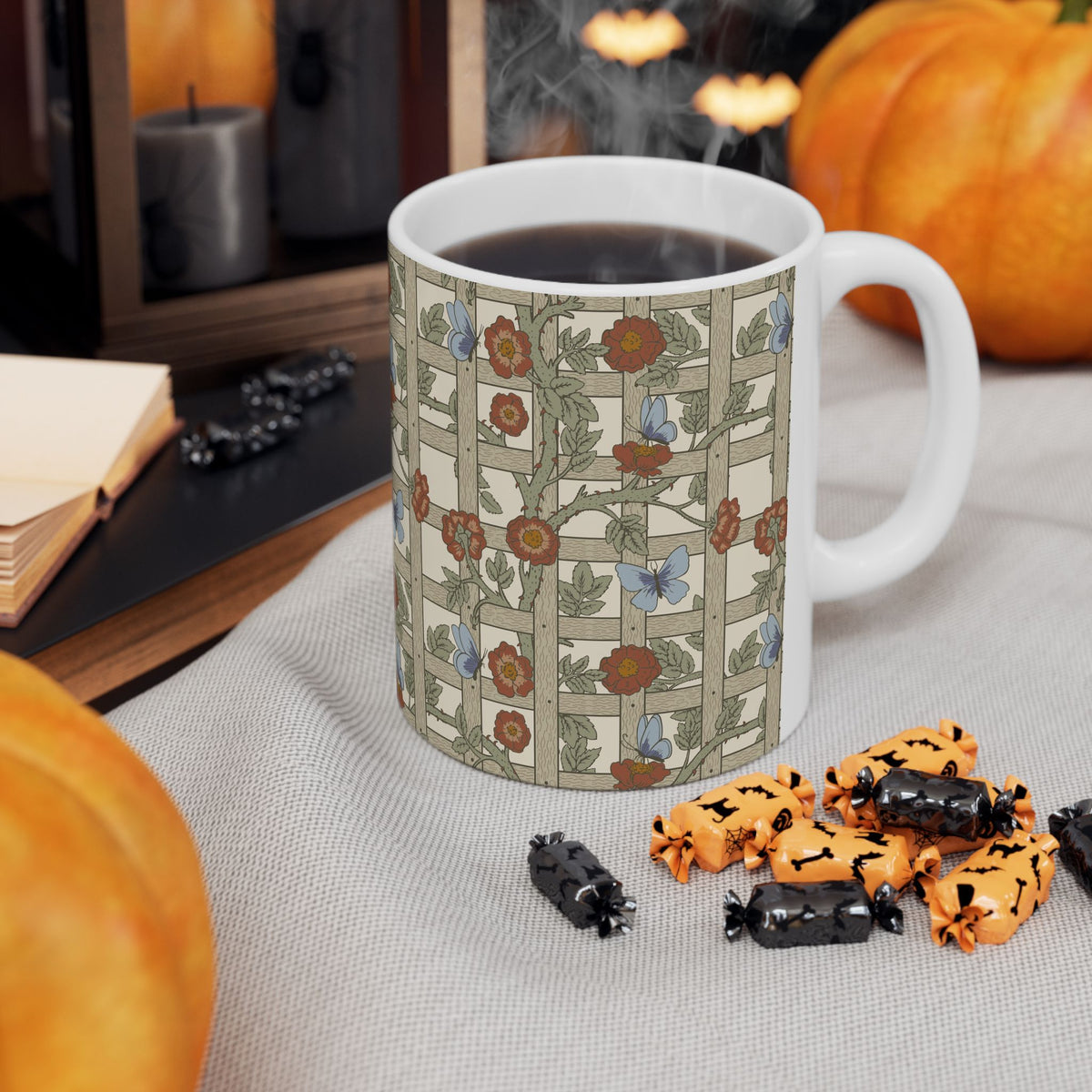 A white ceramic coffee mug with a vintage-style floral and butterfly pattern on a beige grid background, filled with black coffee and set on a light gray fabric tablecloth surrounded by autumn props like pumpkins, candy, and candles.