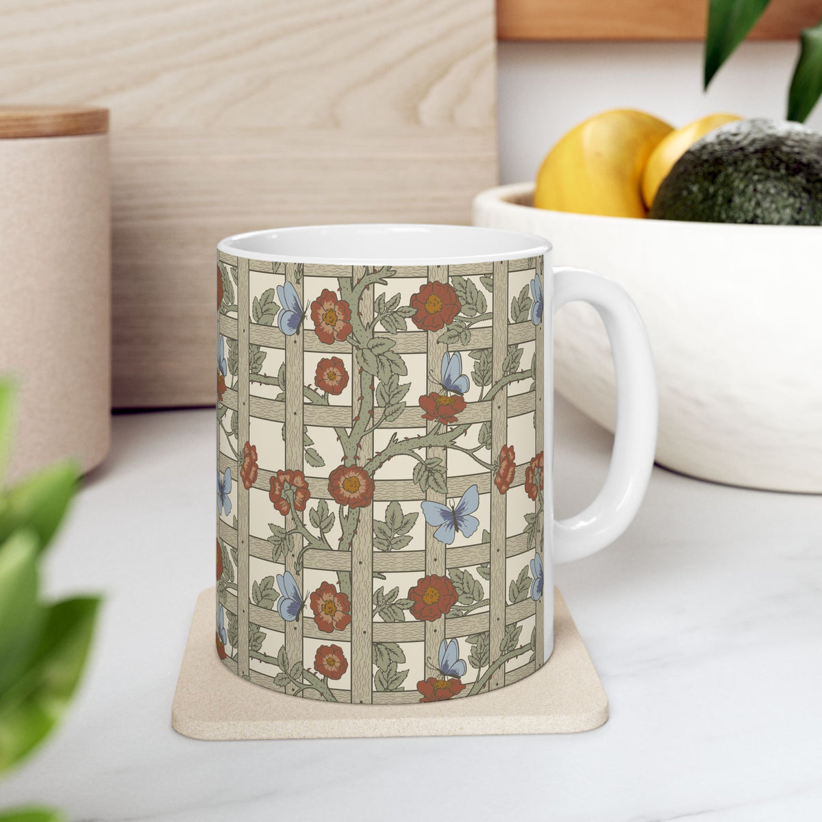 A white ceramic mug with a vintage-style floral trellis pattern, resting on a beige coaster on a white marble countertop with a wooden bowl of fruit in the background.