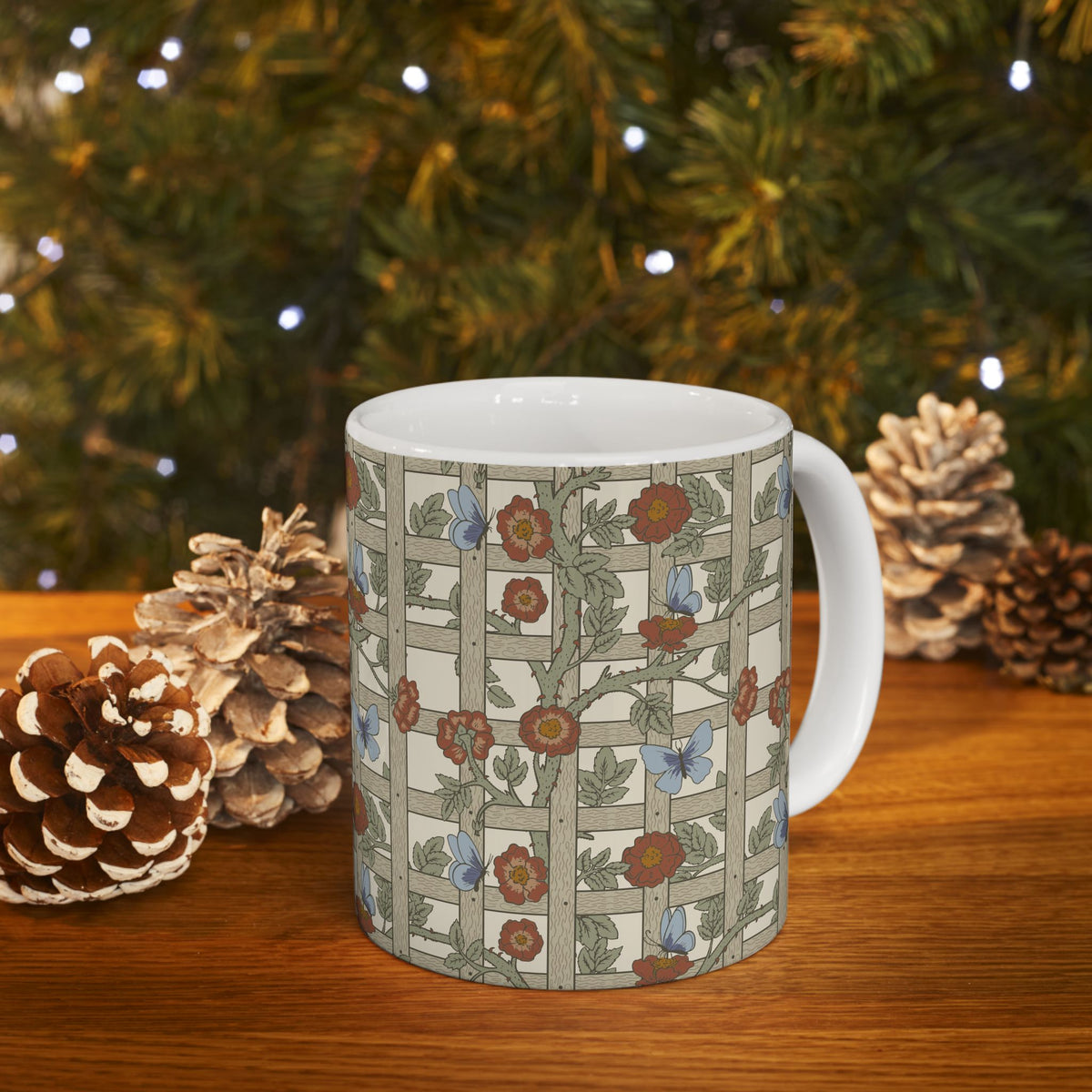 A white ceramic mug with a vintage-style floral and butterfly pattern on a wooden table, surrounded by pinecones and a blurred Christmas tree background with lights.