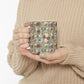 A person in a beige knit sweater holds a ceramic mug with a vintage-style floral and butterfly pattern on a cream background.
