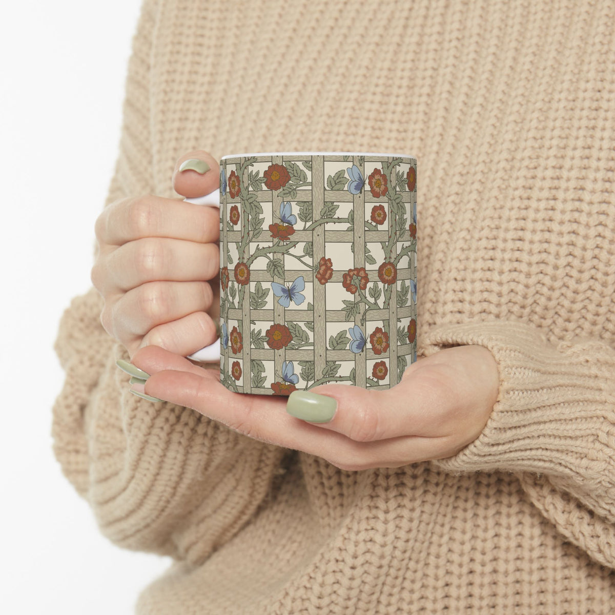 A person in a beige knit sweater holds a ceramic mug with a vintage-style floral and butterfly pattern on a cream background.