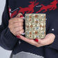 A person in a navy knit sweater with red and white patterns holds a square ceramic mug featuring an Art Nouveau floral vine design on a cream background.