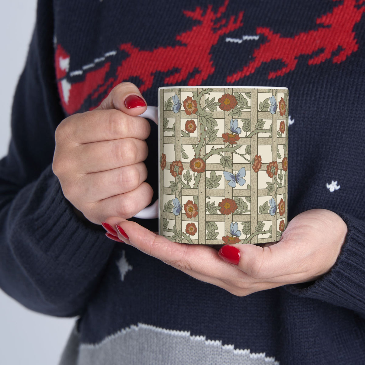 A person in a navy knit sweater with red and white patterns holds a square ceramic mug featuring an Art Nouveau floral vine design on a cream background.