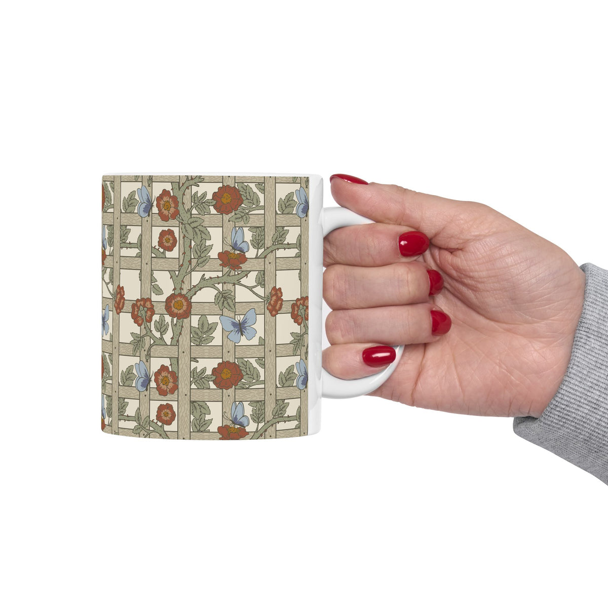 A hand with red nail polish holds a white ceramic mug featuring a vintage-style floral and butterfly pattern in muted green, rust orange, and blue on a beige background.