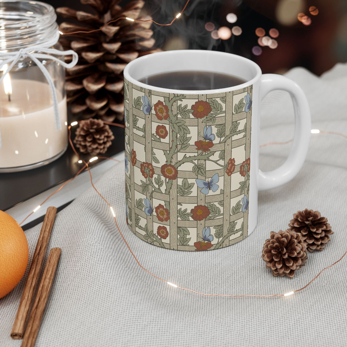 A white ceramic mug with a vintage-style floral and butterfly pattern on a beige grid background, filled with black coffee and set on a light fabric surface with pinecones, a candle jar, cinnamon sticks, and fairy lights.