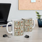Two white ceramic mugs with a vintage Art Nouveau floral pattern, displayed on a light wood desk with a blurred laptop and plant in the background.