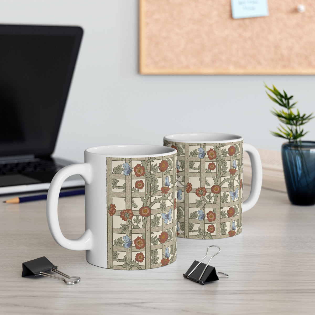 Two white ceramic mugs with a vintage Art Nouveau floral pattern, displayed on a light wood desk with a blurred laptop and plant in the background.
