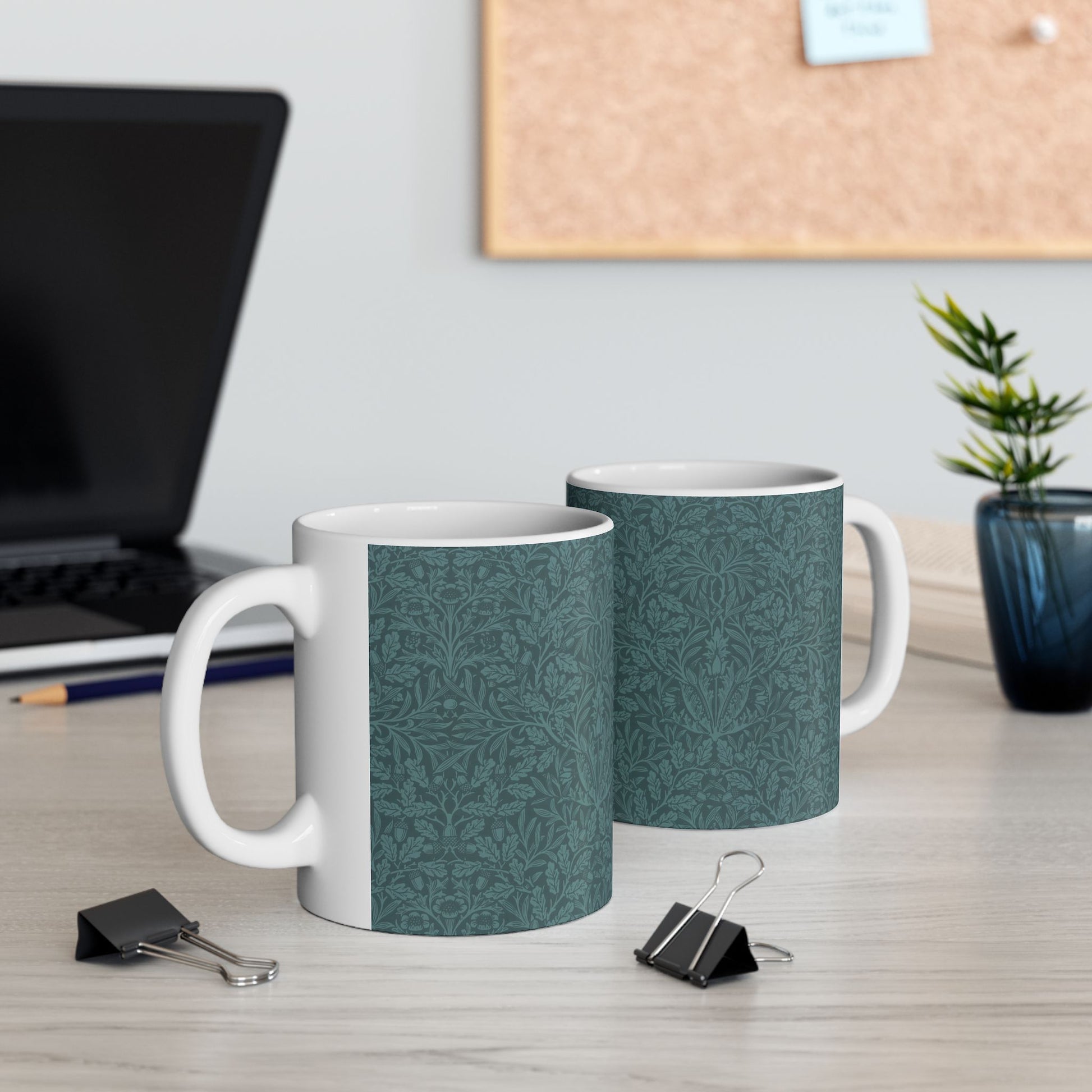 A ceramic mug with a teal base and a white handle featuring a floral and acorn pattern design, inspired by William Morris.