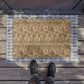 A rectangular doormat with a floral pattern in beige and brown tones, bordered by a blue-gray plaid pattern, shown on a wooden deck with a person's feet in black sneakers standing at the bottom edge.