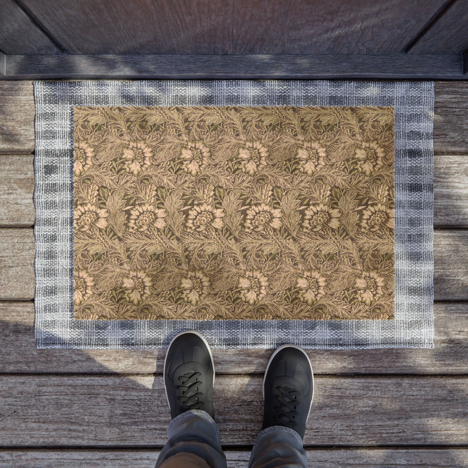A rectangular doormat with a floral pattern in beige and brown tones, bordered by a blue-gray plaid pattern, shown on a wooden deck with a person's feet in black sneakers standing at the bottom edge.