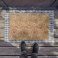 A rectangular decorative doormat with an ornate floral pattern in beige, brown, and muted red, bordered by a blue-gray plaid edge. The mat is placed on weathered wooden deck flooring, with the feet of a person wearing dark sneakers visible at the bottom.