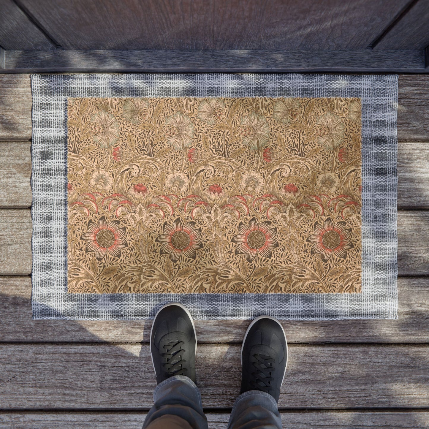 A rectangular decorative doormat with an ornate floral pattern in beige, brown, and muted red, bordered by a blue-gray plaid edge. The mat is placed on weathered wooden deck flooring, with the feet of a person wearing dark sneakers visible at the bottom.