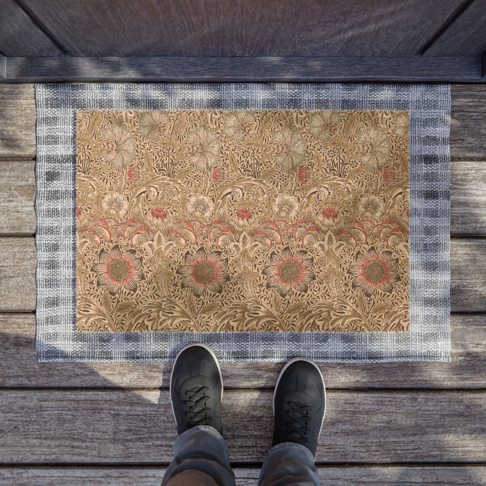 A rectangular decorative doormat with an ornate floral pattern in beige, brown, and muted red, bordered by a blue-gray plaid edge. The mat is placed on weathered wooden deck flooring, with the feet of a person wearing dark sneakers visible at the bottom.