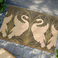 A rectangular doormat featuring a design of two white swans with a green and gold floral background, on a spruce green background.