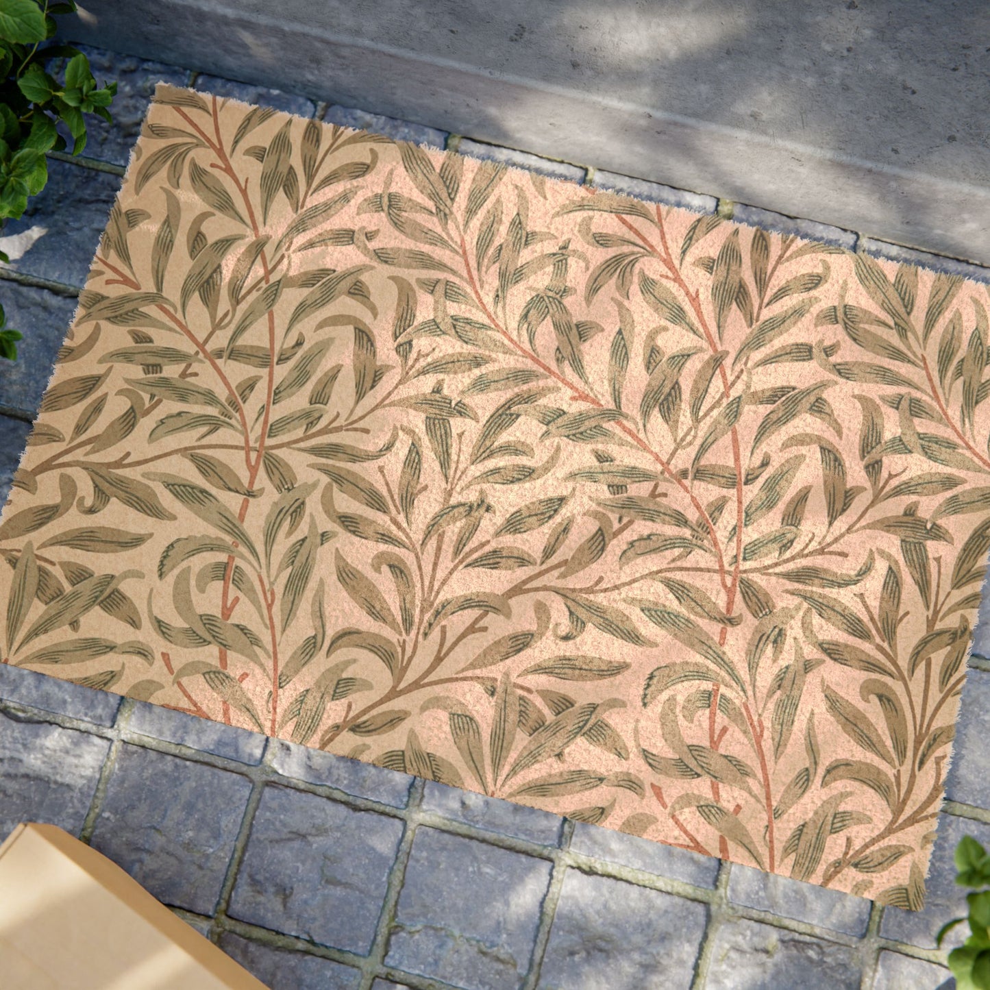 A rectangular area rug with a repeating botanical leaf pattern in muted green and terracotta on a warm beige background, shown placed on gray stone pavers outdoors.