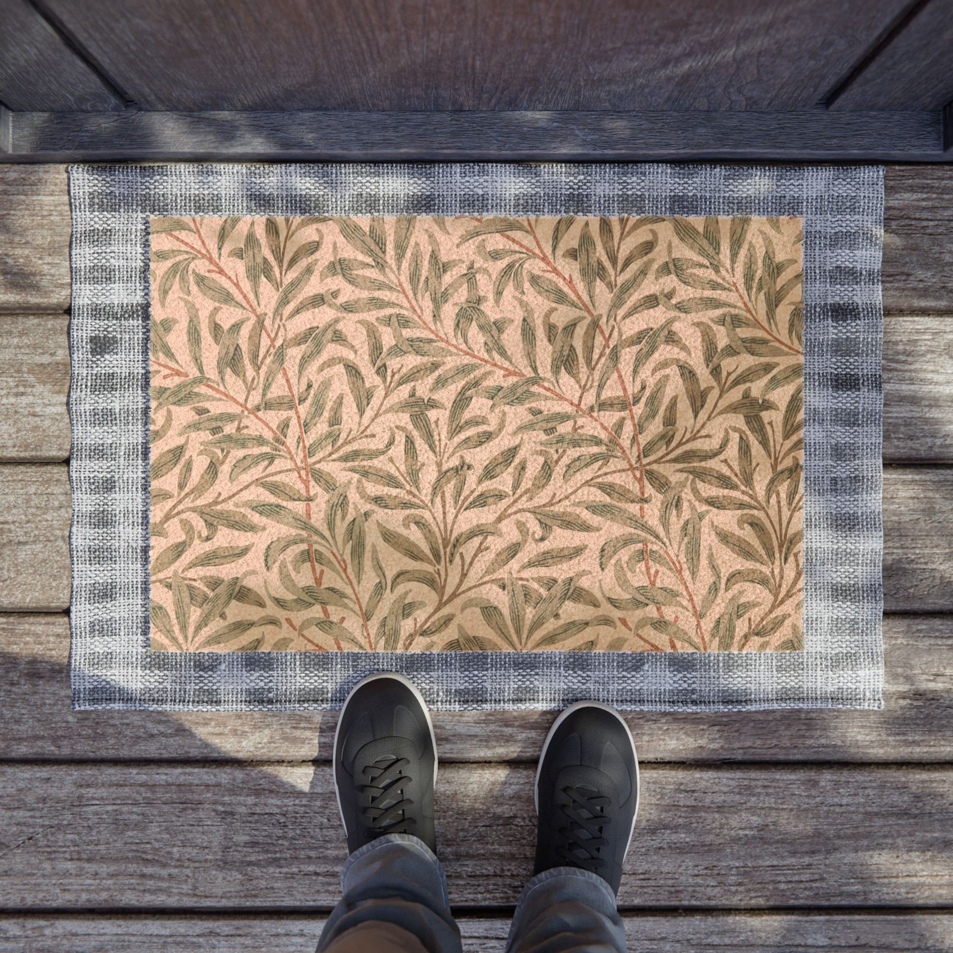 A rectangular doormat with a beige background, green leaf pattern, and gray border, placed on a wooden deck with a person's feet in black sneakers standing at the bottom edge.
