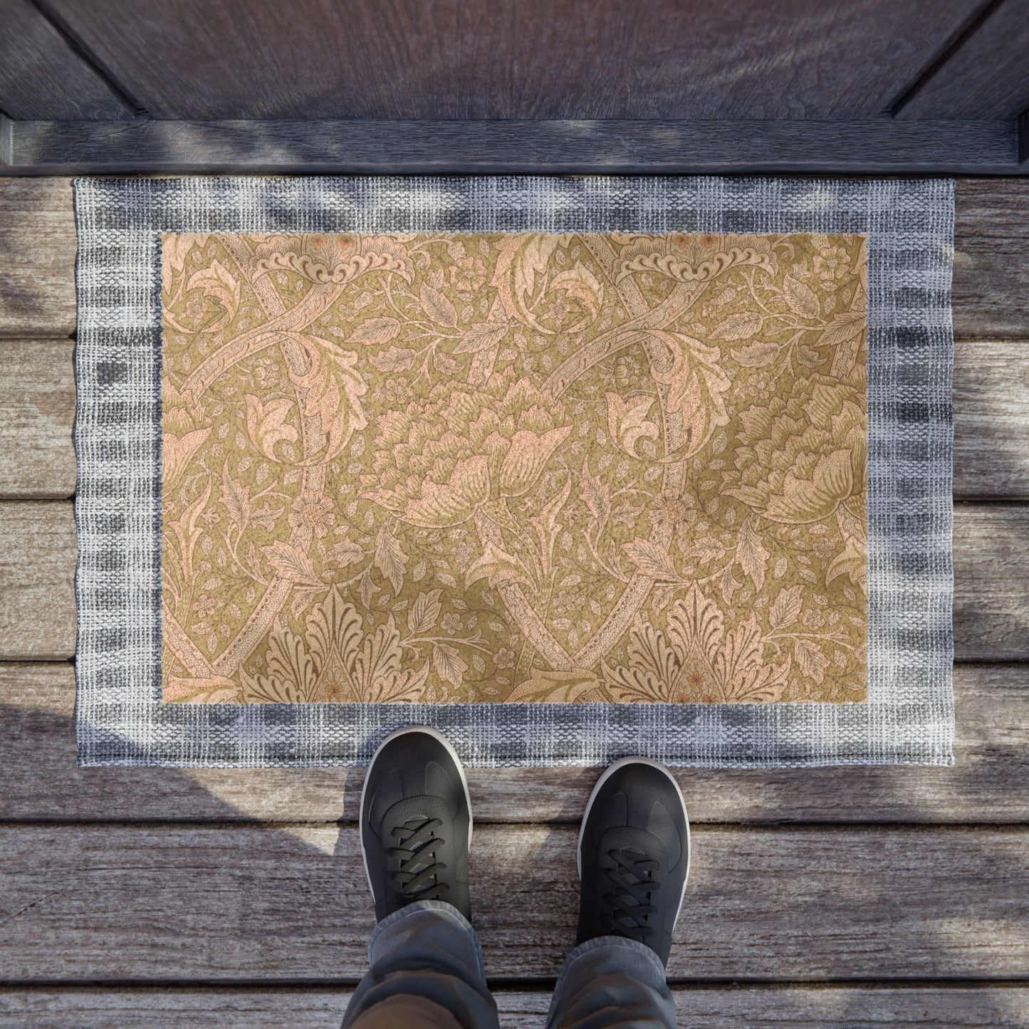 A rectangular doormat with a tan floral damask pattern and a blue-gray plaid border, shown on a wooden deck with a person's feet in dark sneakers standing at the bottom edge.