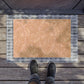 A rectangular doormat with a warm tan floral pattern and a gray plaid border, shown on a wooden deck with a person's shoes visible at the bottom edge.