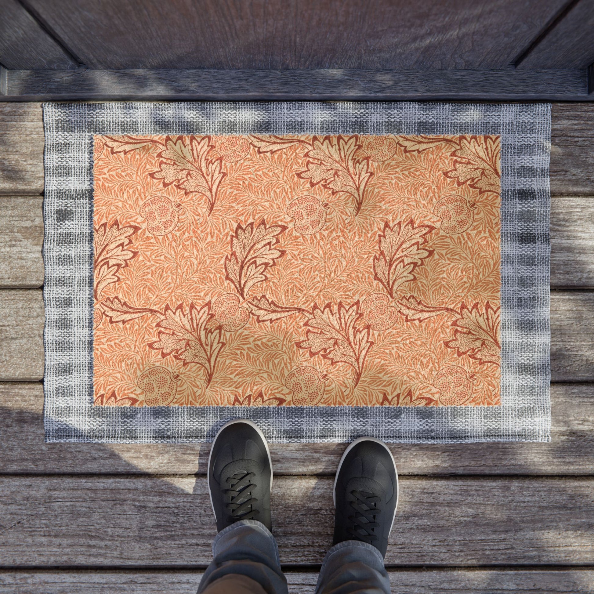A rectangular doormat with a warm peach and tan floral pattern and a gray plaid border, shown on a wooden deck with a person's feet in dark sneakers standing at the bottom edge.
