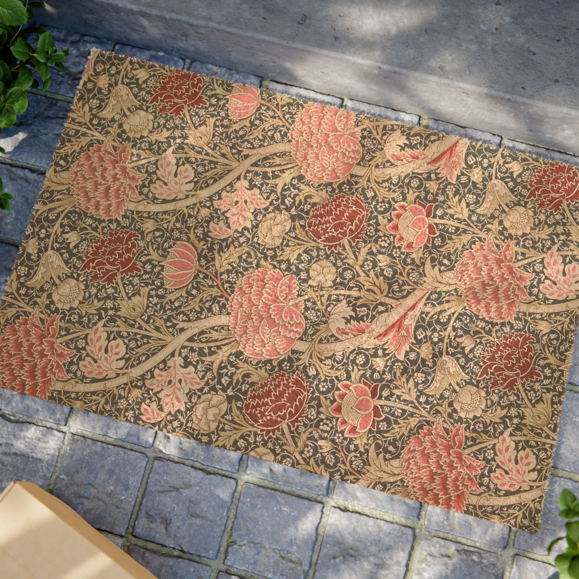 A rectangular woven rug with a dense floral and botanical pattern in muted pink, beige, and brown tones, laid out on gray stone pavers outdoors.