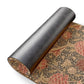 A rolled-up dark gray textured mat or rug shown on a white background, with part of the unrolled section revealing an ornate floral pattern in beige, red, and brown.