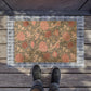A rectangular outdoor doormat with a floral and bird pattern in muted pink, beige, and brown tones, bordered by a gray plaid edge. The mat is placed on weathered wooden deck flooring, with a person's feet in dark sneakers standing at the bottom edge.