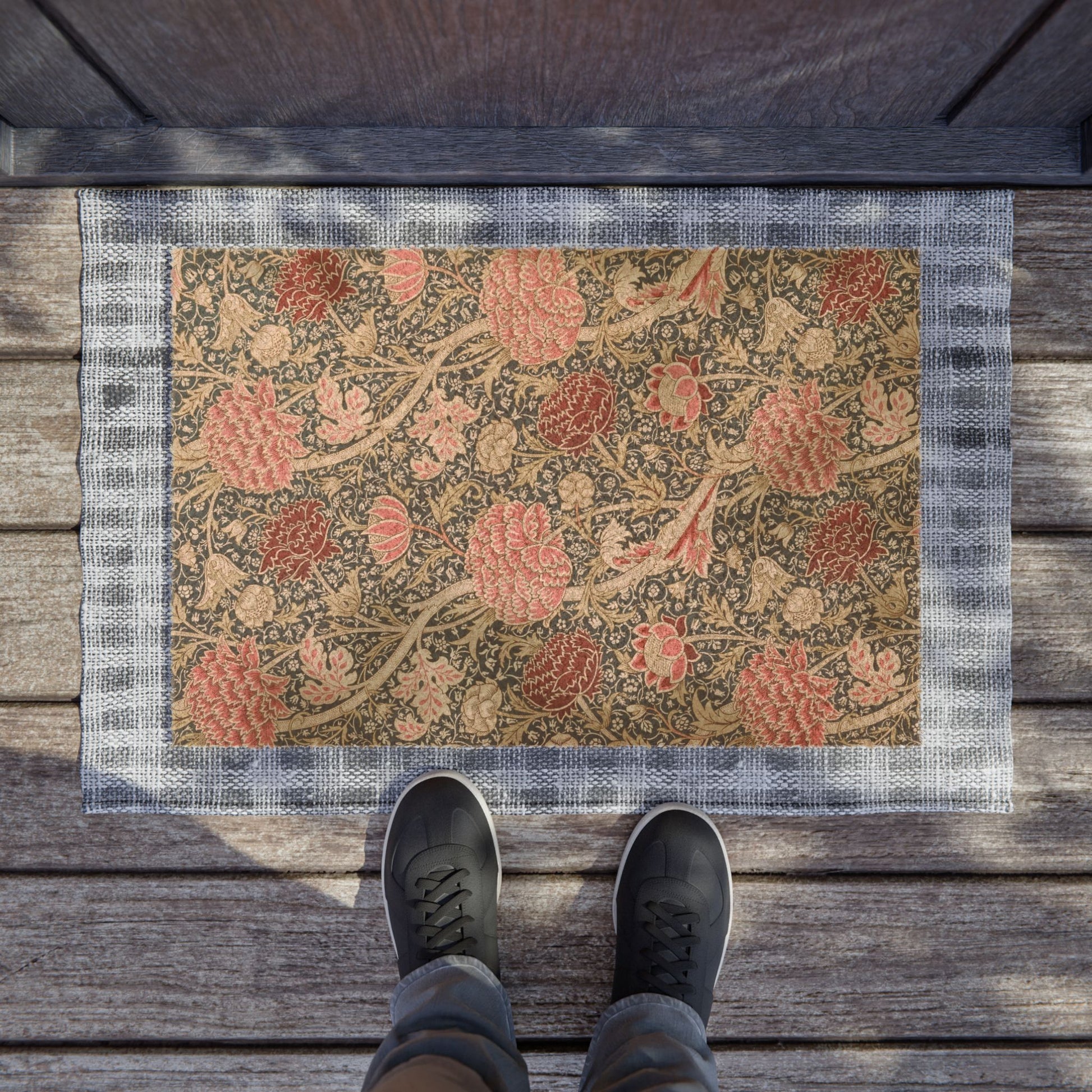 A rectangular outdoor doormat with a floral and bird pattern in muted pink, beige, and brown tones, bordered by a gray plaid edge. The mat is placed on weathered wooden deck flooring, with a person's feet in dark sneakers standing at the bottom edge.