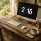 Wooden desk with computer setup, including a monitor displaying '2 16', keyboard, mouse, and a cup of tea.