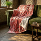 A red and white damask-patterned throw blanket with a soft cream sherpa backing is draped over an olive green velvet sofa in a rustic room with stone floors and warm sunlight.