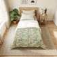 A neatly made bed in a bright, minimalist bedroom. The bedding features a light green and beige floral pattern on the lower half and solid white on the upper half, styled with a light wood headboard, white pillows, a matching wooden nightstand, and a large woven rug on a light wood floor.