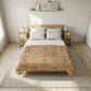 A neatly made bed in a bright bedroom, featuring a floral-patterned quilt in warm beige, pink, and green tones over white bedding and pillows, with a light wood headboard and matching side tables.