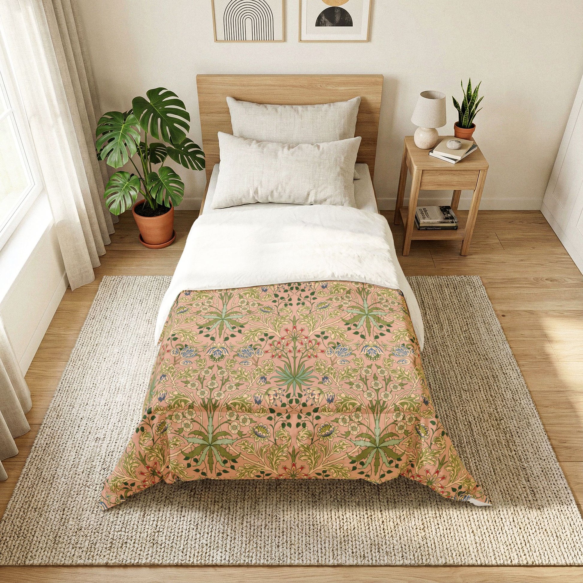 A neatly made single bed in a bright bedroom, featuring a decorative floral and botanical patterned comforter in warm beige, pink, and green tones, with white pillows and bedding. The bed is styled in a cozy room with a wooden headboard, a matching wooden side table, a large potted monstera plant, and a woven rug on a light wood floor.
