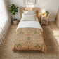 A neatly made bed in a bright bedroom setting, featuring a peach and green botanical patterned comforter with white pillows and a white top sheet. The bed is styled with a light wood headboard, a matching wooden nightstand with a lamp and small plant, and a large woven rug on a light wood floor.