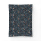 A rectangular fabric item, likely a pillowcase or small blanket, featuring a dense botanical and bird pattern on a dark navy background. The pattern includes light blue leaves, small orange and yellow flowers, and stylized birds in muted pink and brown tones.