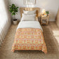 A single bed styled in a bright, modern bedroom, featuring a duvet cover with a warm orange and yellow damask pattern over white bedding. The bed is placed on a textured brown jute rug next to a wooden nightstand with a lit lamp and a potted plant.