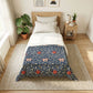 A neatly made bed in a bright, modern bedroom featuring a navy blue floral patterned comforter with red flowers and cream leaves, styled with white sheets and pillows. The bed is placed on a woven jute rug next to a wooden side table with a lamp and books, and a large green plant in a terracotta pot sits near the window.