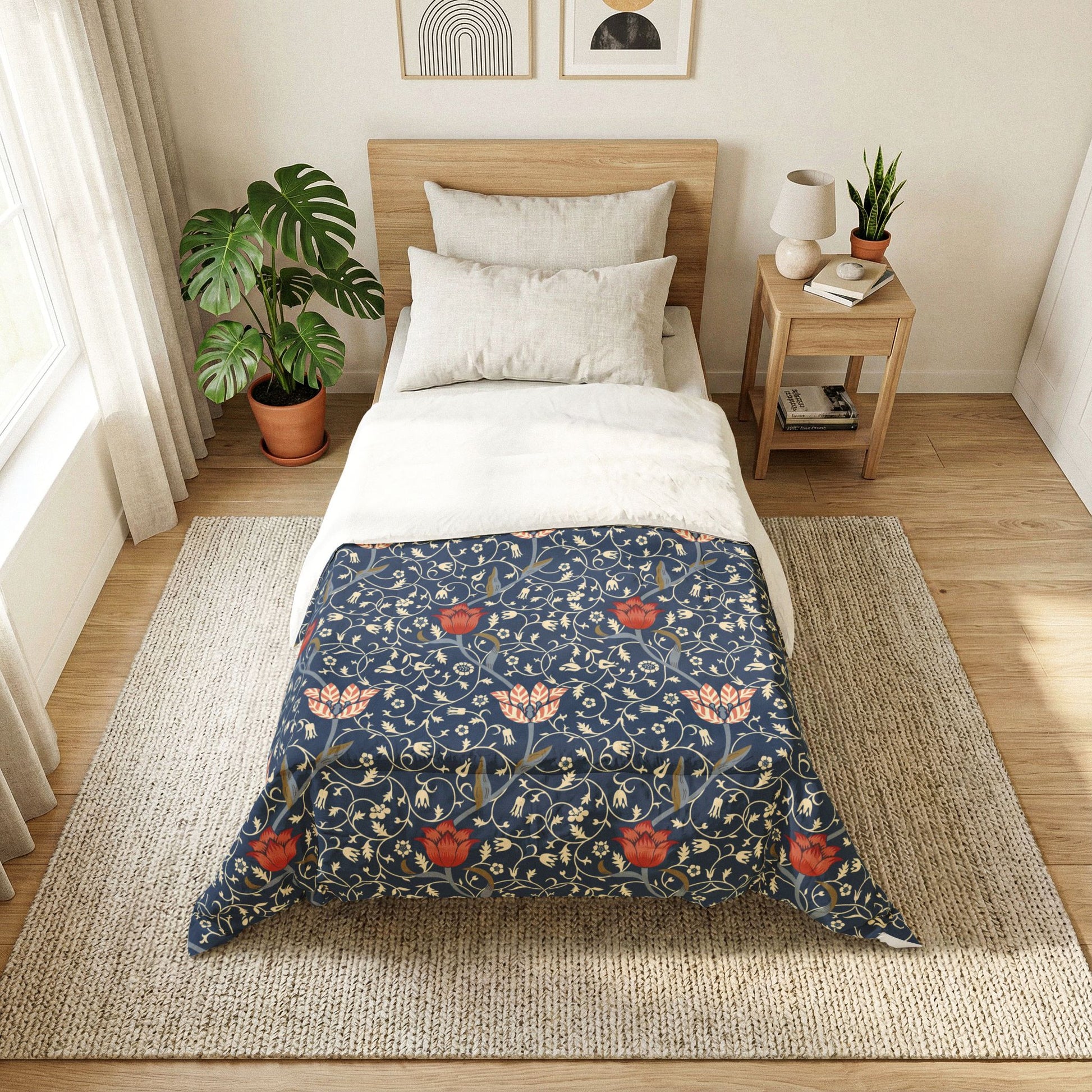 A neatly made bed in a bright, modern bedroom featuring a navy blue floral patterned comforter with red flowers and cream leaves, styled with white sheets and pillows. The bed is placed on a woven jute rug next to a wooden side table with a lamp and books, and a large green plant in a terracotta pot sits near the window.