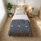 A single bed styled in a bright, modern bedroom, featuring a navy blue comforter with a red and pink floral pattern, layered over white sheets and pillows. The bed is set on a woven jute rug next to a wooden nightstand with a lamp and a potted plant.