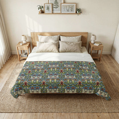 A bed dressed in a white top sheet and a colorful patterned quilt or comforter, styled in a bright, minimalist bedroom with wooden nightstands, lamps, and framed botanical prints on the wall.