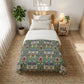A neatly made bed in a bright bedroom setting, featuring a decorative patterned comforter with a blue, green, and red botanical design over white bedding. The bed is styled with a light wood headboard, a matching side table with a lamp and small plant, and a large woven rug on the floor.