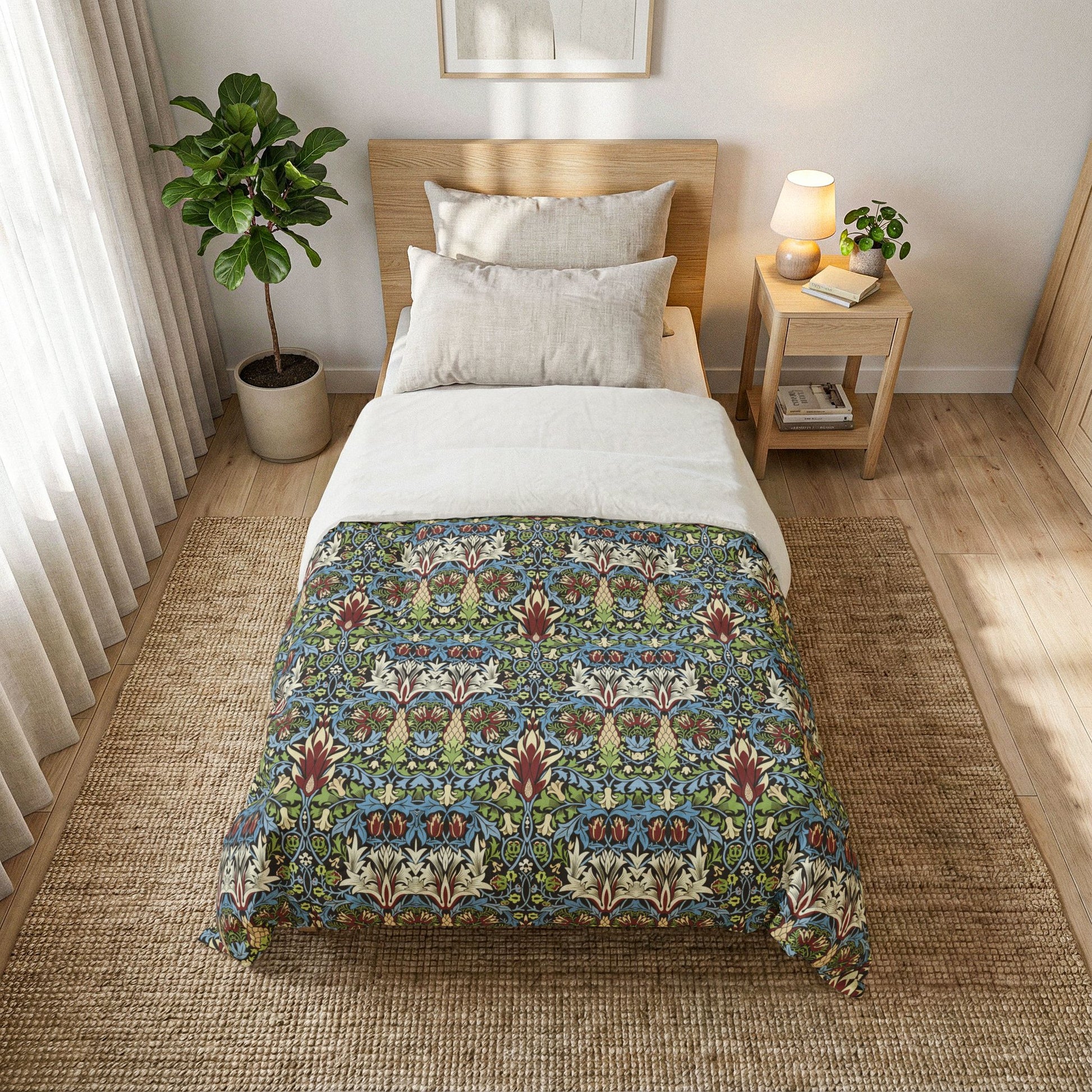 A neatly made bed in a bright bedroom setting, featuring a decorative patterned comforter with a blue, green, and red botanical design over white bedding. The bed is styled with a light wood headboard, a matching side table with a lamp and small plant, and a large woven rug on the floor.