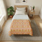 A neatly made bed in a bright bedroom, featuring a white duvet cover with a prominent orange and yellow ornamental pattern on the lower half. The bed is styled with a light wood headboard, white pillows, a beige rug underneath, and a small wooden side table with a lamp and books.