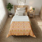 A neatly made bed in a bright bedroom, featuring a patterned comforter in orange, yellow, and green tones. The room includes a wooden headboard, neutral pillows, a bedside table with a lamp, a potted plant, and a woven rug.