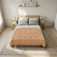 A bed dressed in a patterned orange and yellow damask-style comforter or quilt, styled in a bright, modern bedroom with light wood furniture, neutral walls, and a textured rug.