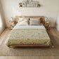 A styled bedroom scene showing a bed made with a light beige floral and bird-patterned quilt or comforter, white sheets, and neutral pillows. The bed is placed on a woven rug in a bright, neutral-toned room with wooden nightstands, lamps, and wall art.