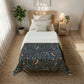 A neatly made bed in a bright bedroom, featuring a dark navy patterned comforter with birds and floral motifs, layered over white bedding. The room includes a wooden headboard, a bedside table with a lamp, a potted plant, and a woven rug.